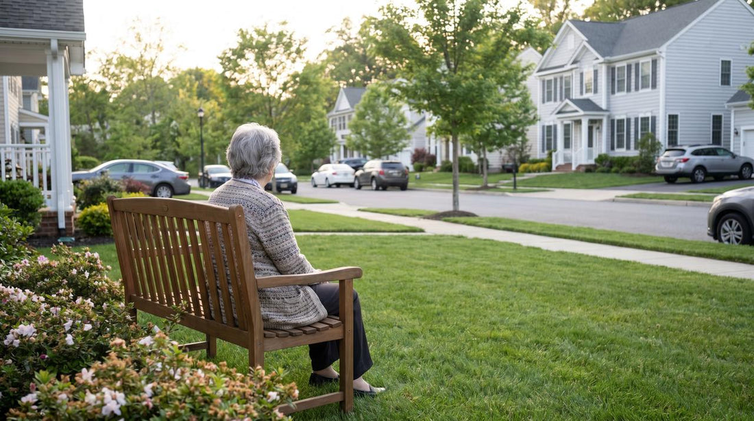 An elderly woman with grey hair viewed from behind, sitting alone on a wooden bench in a green front yard, looking out toward a quiet suburban street lined with houses and cars.